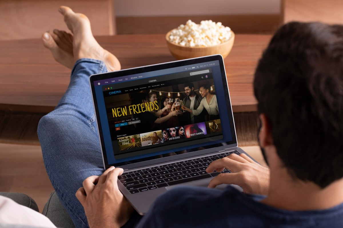 Ein Mann sitzt bequem auf einem Sofa mit übergeschlagenen Beinen und schaut auf seinem Laptop eine Streaming-Plattform mit einem Film namens „New Friends“. Neben ihm steht eine Schüssel Popcorn, und er genießt die Inhalte, die er mit seiner Kreditkarte abonniert hat.