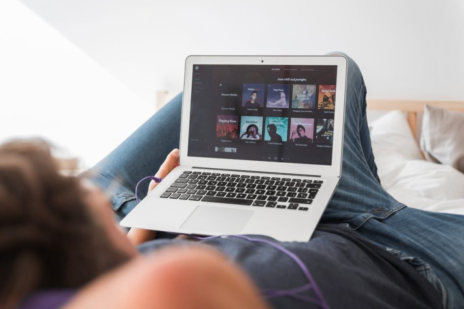 Ein junger Mann liegt entspannt auf einem Bett und hält ein modernes Laptop auf seinem Schoß, auf dessen Bildschirm die Spotify-Oberfläche mit verschiedenen Musik-Playlisten angezeigt wird. Er hört Musik über ein Abo, das er mit einer seiner Kreditkarten bezahlt hat. Das Licht ist weich, und die Szene vermittelt eine ruhige, gemütliche Atmosphäre beim Musikhören.