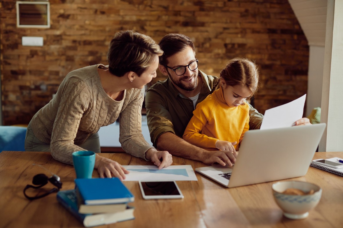 Ein Vater sitzt lächelnd mit seiner Tochter vor einem Laptop, während die Mutter sich zu ihnen beugt und gemeinsam Unterlagen durchgeht. Das warme Licht und die gemütliche Atmosphäre zeigen eine Familie, die Zeit miteinander verbringt und zugleich über Finanzen oder Zukunftsplanung spricht